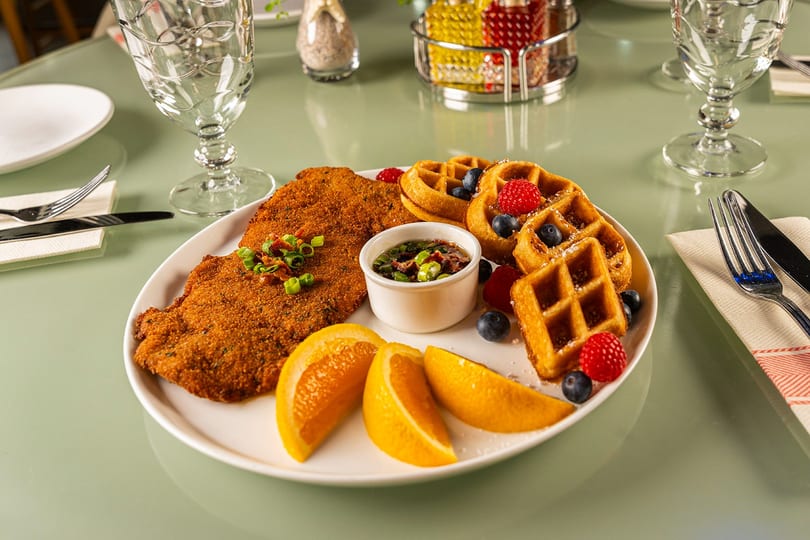 Crispy breaded cutlet, mini waffles with berries, orange slices, and sauce on a green table.