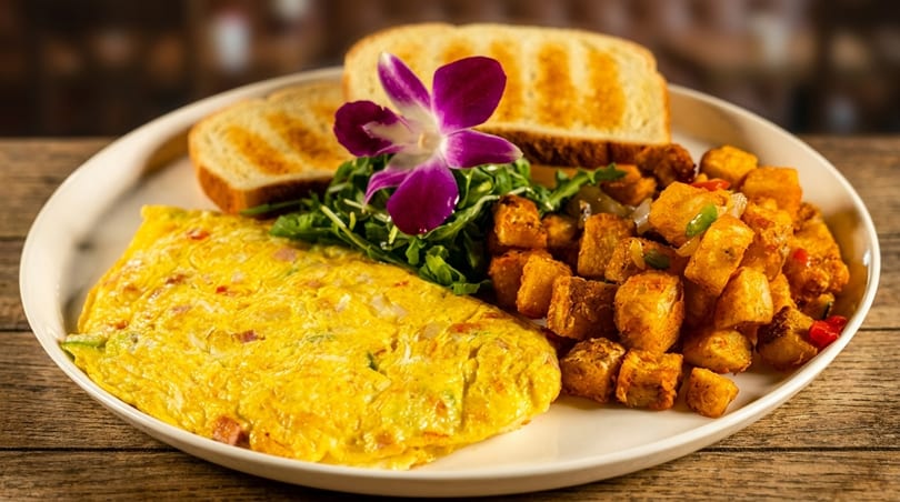 A breakfast plate with a fluffy omelet, crispy home fries, toast, and a purple orchid garnish.