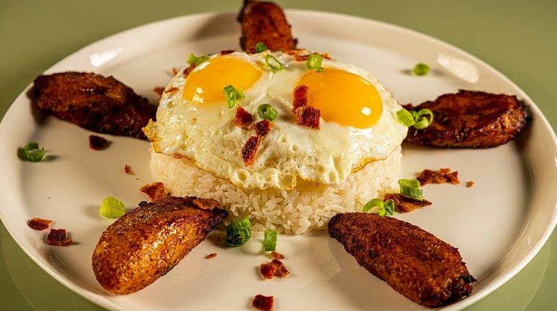 White rice topped with two fried eggs, bacon bits, and green onions, surrounded by fried plantains.