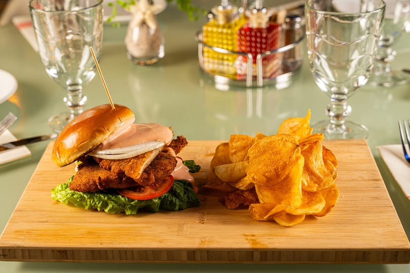 Breaded sandwich with sauce, onions, lettuce, and tomato, served with chips on a wooden board.