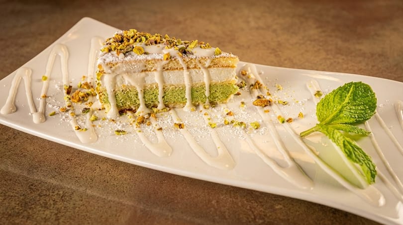 A slice of pistachio cake with white frosting, nuts, and a mint leaf on a white plate.