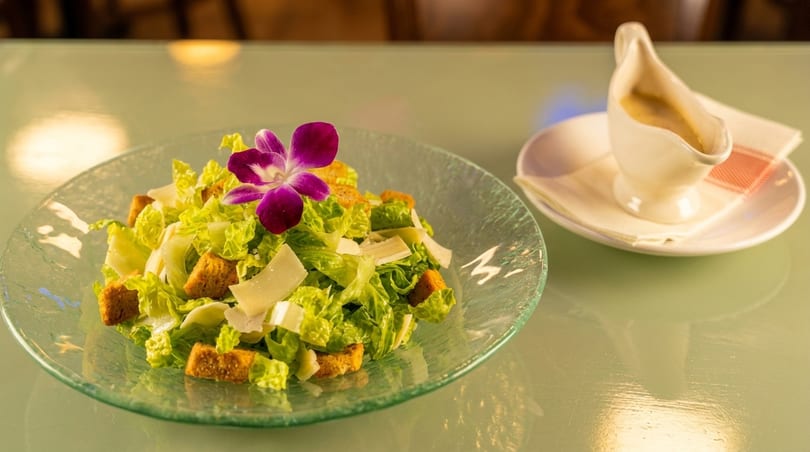 Caesar salad with croutons, parmesan, and a purple orchid, with a side of dressing.