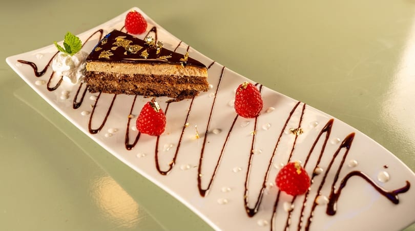 A slice of chocolate cake with gold flakes, whipped cream, mint, and raspberries on a white plate.