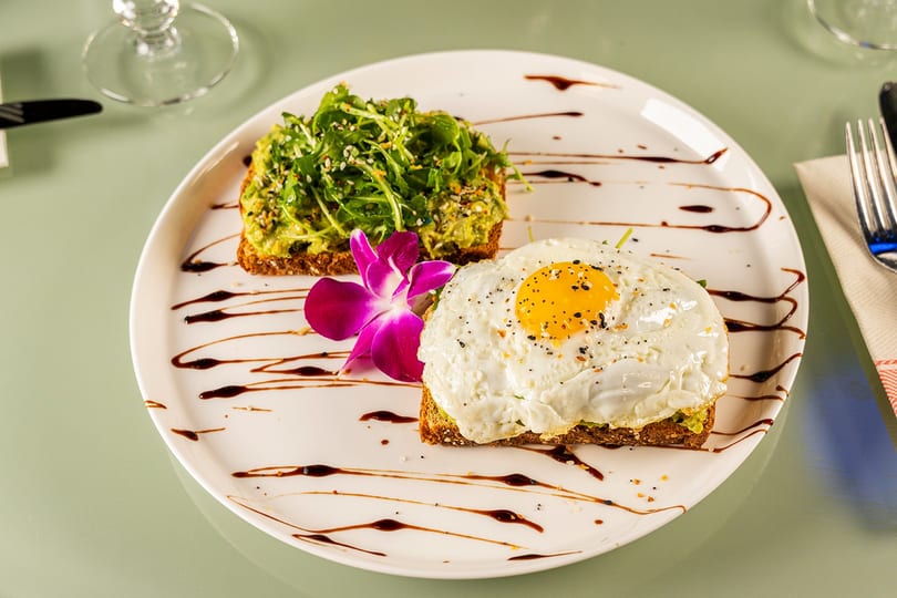Avocado toast with arugula, a fried egg, and a purple orchid garnish on a white plate.