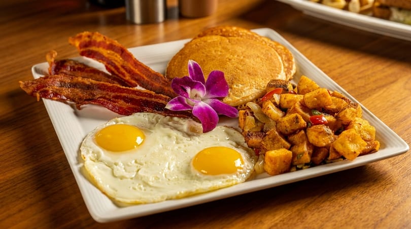 A breakfast plate with bacon, fried eggs, pancakes, and home fries, garnished with a purple orchid.