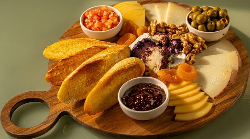 A wooden board with sliced bread, cheeses, olives, nuts, dried apricots, and dips.