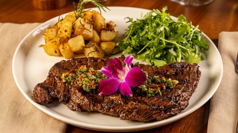 Grilled steak with chimichurri, a purple orchid, roasted potatoes, and arugula on a white plate.