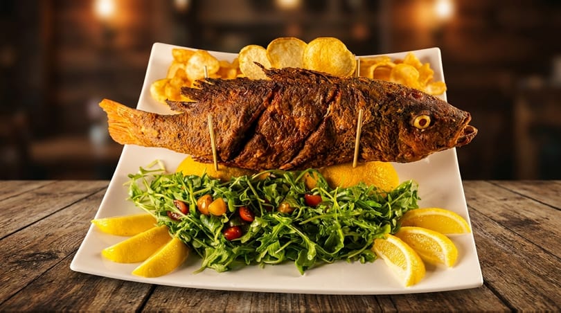 A whole fried fish with lemon wedges, arugula salad, and potato chips on a white platter.